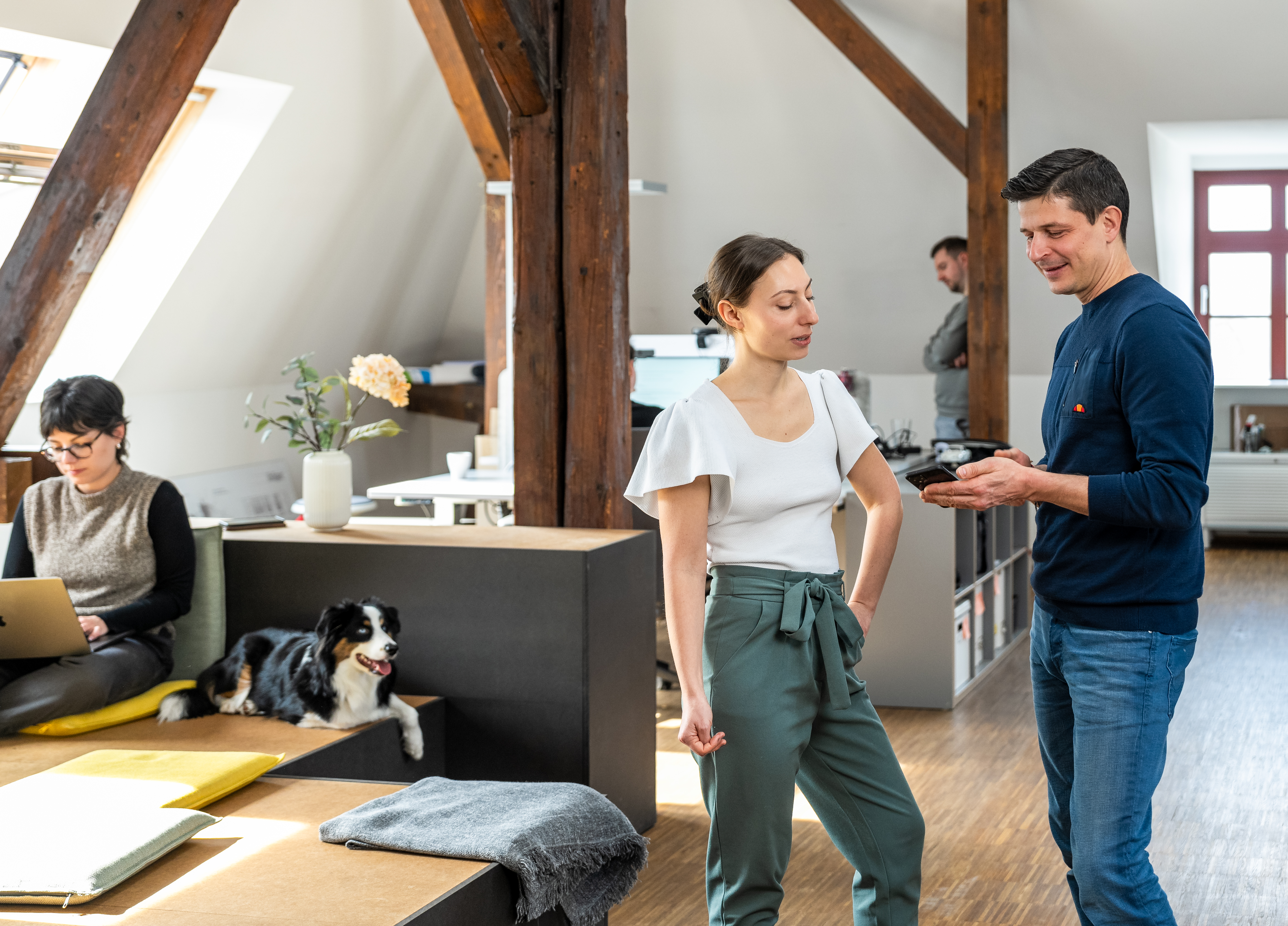 Mehrere Personen in einem UXMA-Büro. Im Vordergrund stehen eine Frau und ein Mann und unterhalten sich, während der Mann etwas an seinem Smartphone zeigt.