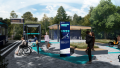 Eine moderne Mobilitätsstation im Freien mit Leih-E-Bikes und E-Scootern, einer Bushaltestelle mit digitaler Anzeige sowie überdachten Wartebereichen. Mehrere Menschen bewegen sich durch die Station, die von Bäumen und blühenden Sträuchern umgeben ist.