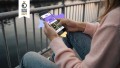 Person in rosanem Hoodie und Jeas hält in ihrer rechten Hand ein Smartphone mit der Startseite von „JUP!“. Links oben ist das Logo vom German Design Award Winner 2027 platziert.
