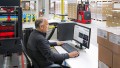 Ein älterer Mann sitzt vor einem Monitor und betrachtet die Startseite des Kundenportals für Linde Material Handling. Er befindet sich in einer großen Lagerhalle und ist umgeben von Paletten voller Pakete. Zudem sind zwei Gabelstapler zu sehen.