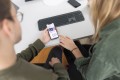 Man sieht eine Büroszene in der zwei Personen von oben auf die Oberfläche eines Smartphones schauen und diese disktuieren.