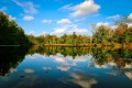 Eine herbstliche Landschaft mit Wasser im Vordergrund und Bäumen im Hintergrund. Der Himmel ist blau und wolkig.