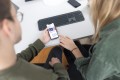 Ein Mann hält ein Handy in der Hand auf dem Website-Inhalte abgebildet sind und eine, die rechts daneben sitzt, Frau zeigt auf diese Inhalte.