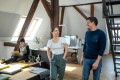 Ein Mann und eine Frau unterhalten sich im Gehen. Die Frau trägt ein weißes T-Shirt mit Flügelärmeln und dazu eine olivgrüne Stoffhose mit geschnürtem Gürtel. Der Mann trägt eine Jeans und einen dunkelblauen Strickpullover. Im Hintergrund erkennt man eine offene Büroumgebung mit freigelegten Holzbalken. Im hinteren Bereich des Raumes stehen Schreibtische an denen Personen arbeiten. In der linken Seite des Bildes sitzt eine Frau mit Laptop auf ihren Schoß, neben ihr liegt ein Hund.