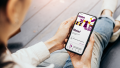 A seated person holds a smartphone in his hand, on which the home screen of the “Moin CAU” app is open. In addition to a vector-based illustration in yellow, purple and black and a greeting, a tile with current messages and a tile with to-dos are visible. At the bottom of the screen there is a navigation bar with four icons.