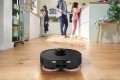 A family in a kitchen in the background. The BOSCH vacuuming and mopping robot in the foreground, cleaning up dirt and grime from kitchen chores.