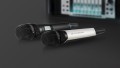 Two radio microphones, one completely black, one white-silver, lie one behind the other on a black background. The rear black microphone is switched on. The number of megahertz can be read on the integrated screen.