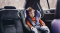 A small child sleeps relaxed in a child seat. Speakers are sketched to the right and left of his head.