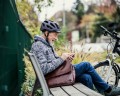 Ein älterer Mann mit Fahrradhelm sitzt auf einer Bank. Er hält ein Smartphone in der Hand und tippt begeistert darauf herum. Vor ihm steht ein Fahrrad.