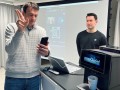 Two people stand in front of a screen. One of them is holding a cell phone in one hand, with the other he performs a gesture that is reflected in an interface that is projected onto a screen in the background.