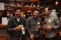 Three men standing at a bar, all holding a drink in their hands and laughing joyfully.