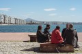 Five people are sitting in the sun on a bench on a promenade. In the background you can see the white tower, a landmark of Thessaloniki.