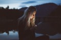 A blonde woman sits by a lake at dusk. Her face is illuminated only by the smartphone she is looking at.