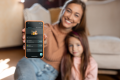 A smiling mother holds a smartphone with the Carbster app opened towards the viewer. Next to her, her little daughter leans in her arms.